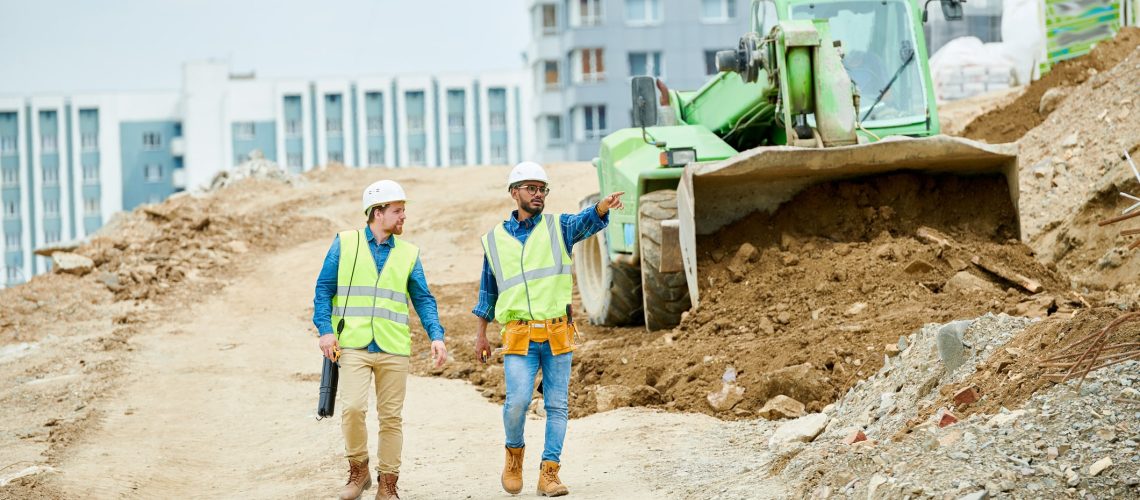 Two guys in helmets and waistcoats walking near digger and pointing at distance during inspection on construction site