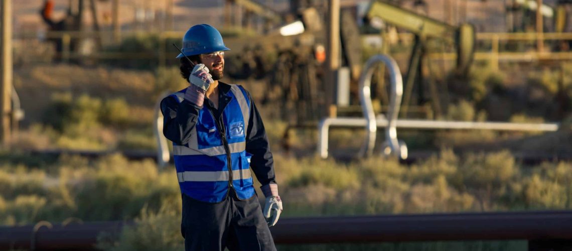 A medium wide shot of an Indian man in a blue hi-vis and helmet using a walkie-talkie in an oilfield on a sunny day in Kern County, California.